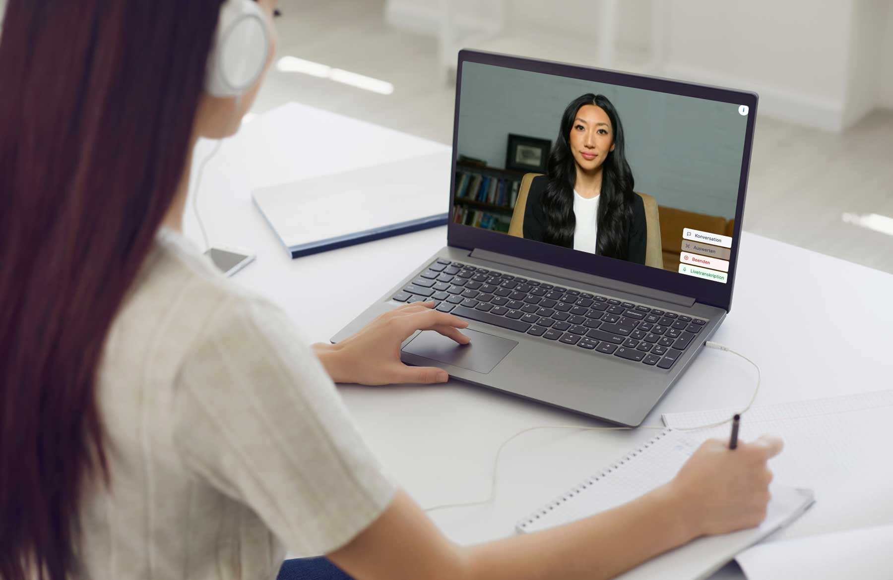 Eine Pharmaziestudentin mit Kopfhörern sitzt am Laptop und unterhält sich mit einem Avatar auf dem Bildschirm, während sie nebenher in einem Notizblock schreibt.